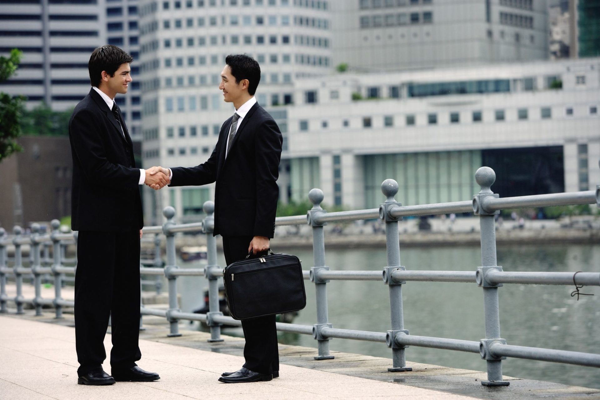 Two,Businessmen,Shaking,Hands,,Buildings,In,The,Background